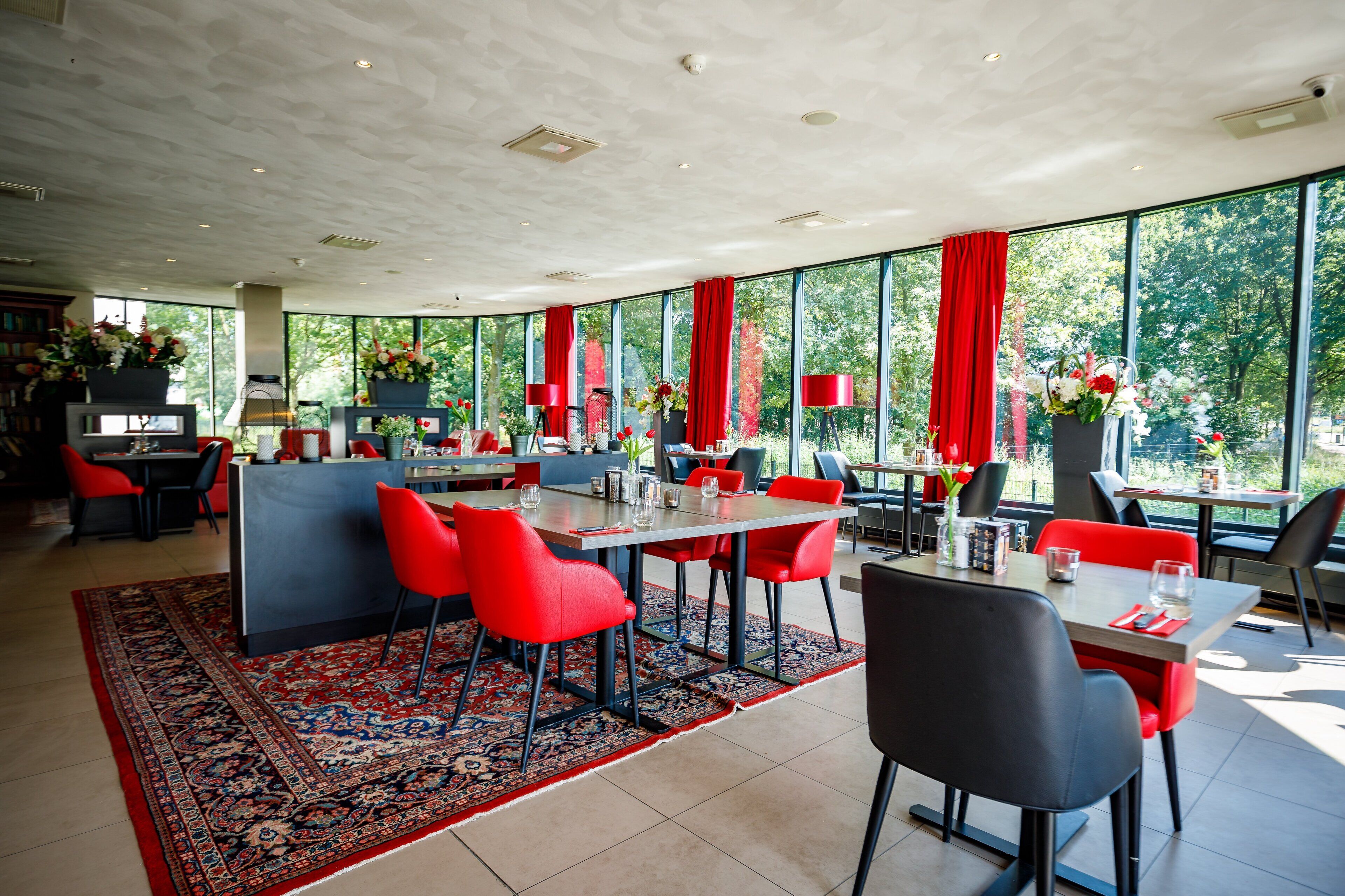 Een modern restaurant met grote ramen, rode stoelen en een kamerbreed tapijt.