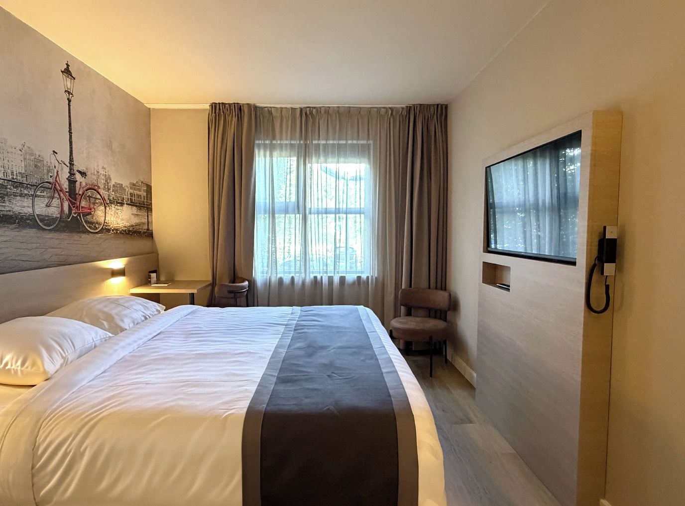 A hotel room with a bed, a chair, a desk, a lamp, a painting, and a window with curtains.