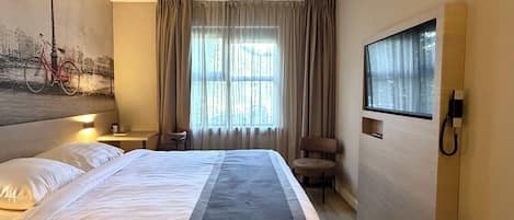 A hotel room with a bed, a chair, a desk, a lamp, a painting, and a window with curtains.
