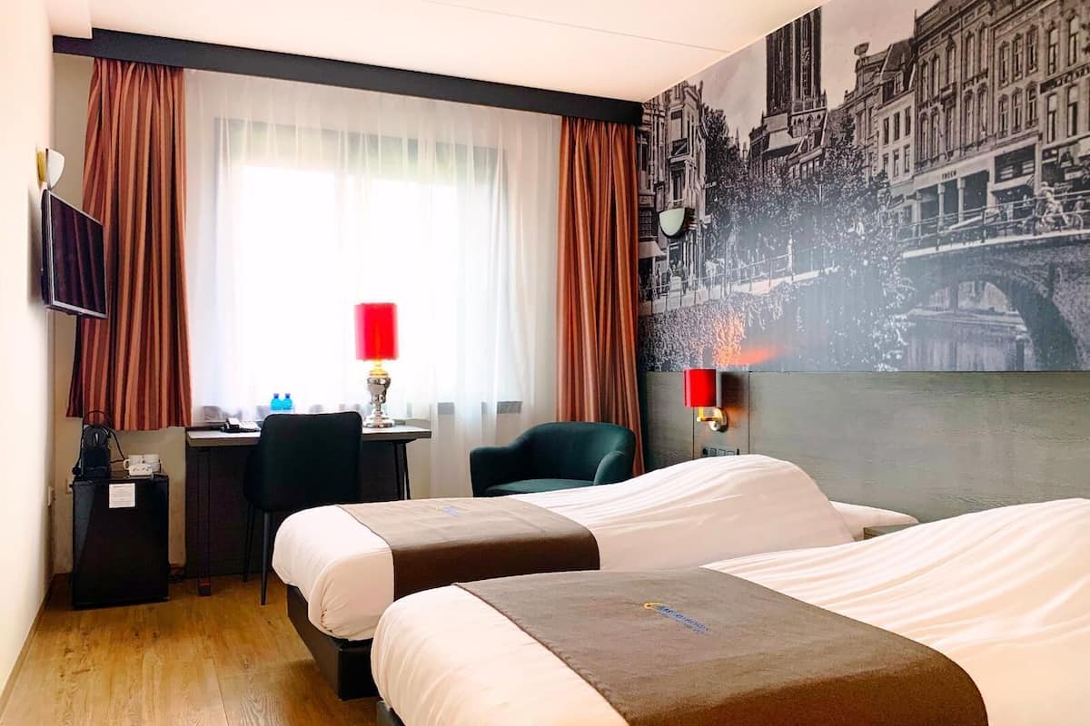 Une chambre d’hôtel avec deux lits, un bureau, une chaise, un téléviseur et une grande fresque murale représentant un paysage urbain.