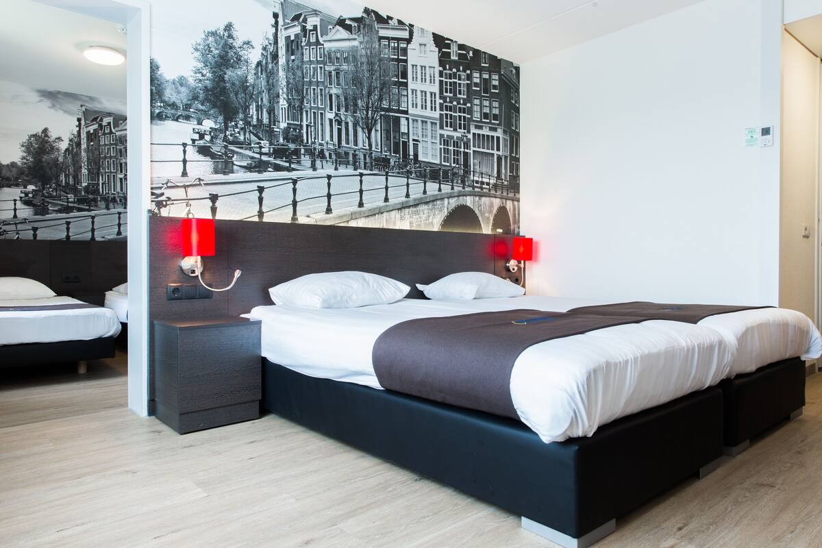 A modern hotel room with a large bed, wooden flooring, a cityscape mural on the wall, and two bedside lamps.