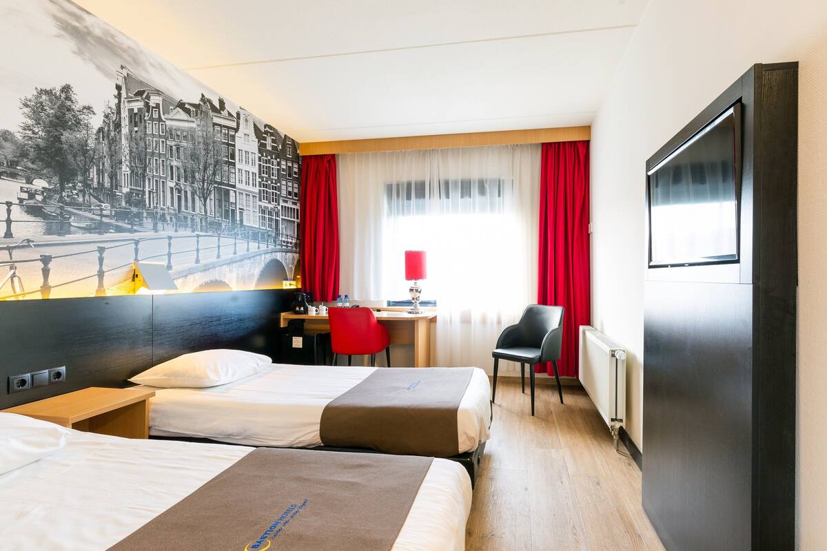 A hotel room with two beds, a desk, a chair, a TV, and a mural of a cityscape.