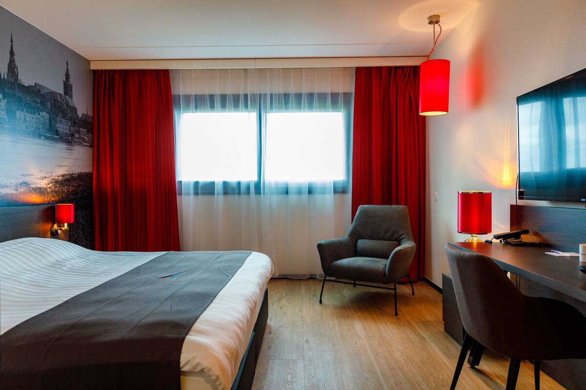 A hotel room with a bed, two chairs, a desk, a television, and a window with red curtains.