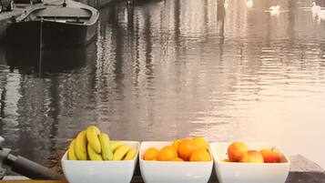 Vue d’un canal avec des bateaux et le reflet d’un bâtiment. Au premier plan, on aperçoit trois bols blancs contenant des bananes, des oranges et des pommes.