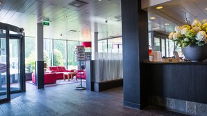 A modern lobby with a reception desk, red seating area, and a large potted plant. - Bastion Hotel Amsterdam Amstel (Amsterdam-Duivendrecht)