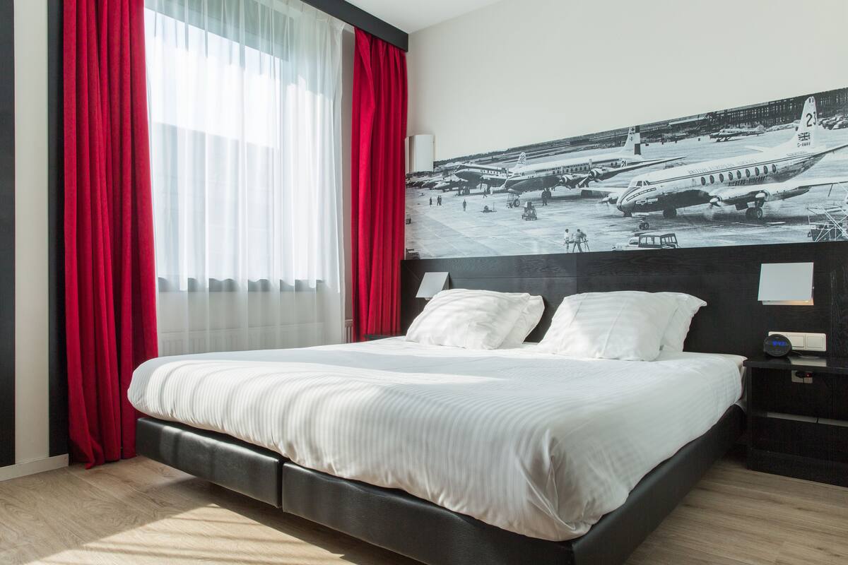 Une chambre d’hôtel avec un grand lit, des rideaux rouges, une table de chevet et une horloge fixée au mur.