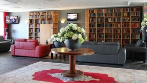 A spacious lobby with a red sofa, grey sofa, and a round table with a vase of flowers. There are multiple bookshelves filled with books and a television mounted on the wall. - Best Western Plus Amsterdam Airport Hotel (Hoofddorp)