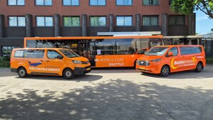 Zwei orangefarbene Shuttlebusse vor einem Gebäude mit Backsteinfassade und großen Fenstern.