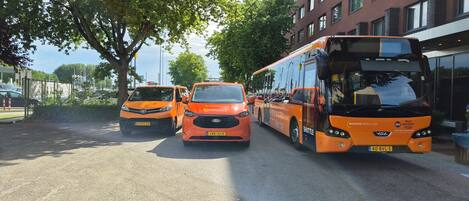 A row of orange shuttle buses parked on a street with a building in the background.
