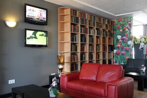 A living room with a red leather sofa, a bookshelf filled with books, two wall-mounted TVs, and a floral curtain.