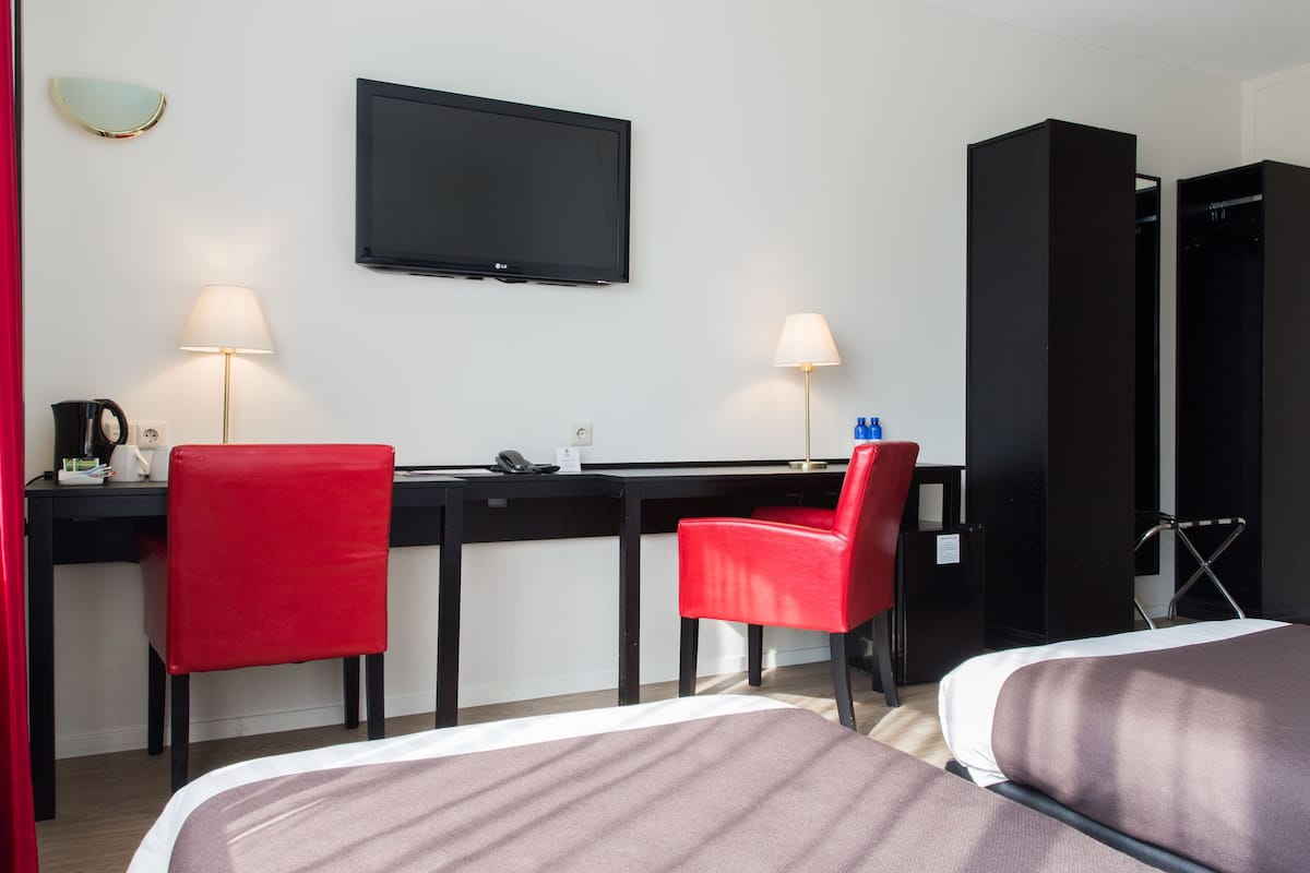 Une chambre d’hôtel avec un bureau, deux chaises rouges, un lit, une télévision et deux lampes.