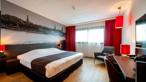 A hotel room with a large bed, a desk, a chair, and a view of a cityscape through the window. - Best Western Plus Amsterdam Airport Hotel (Hoofddorp)