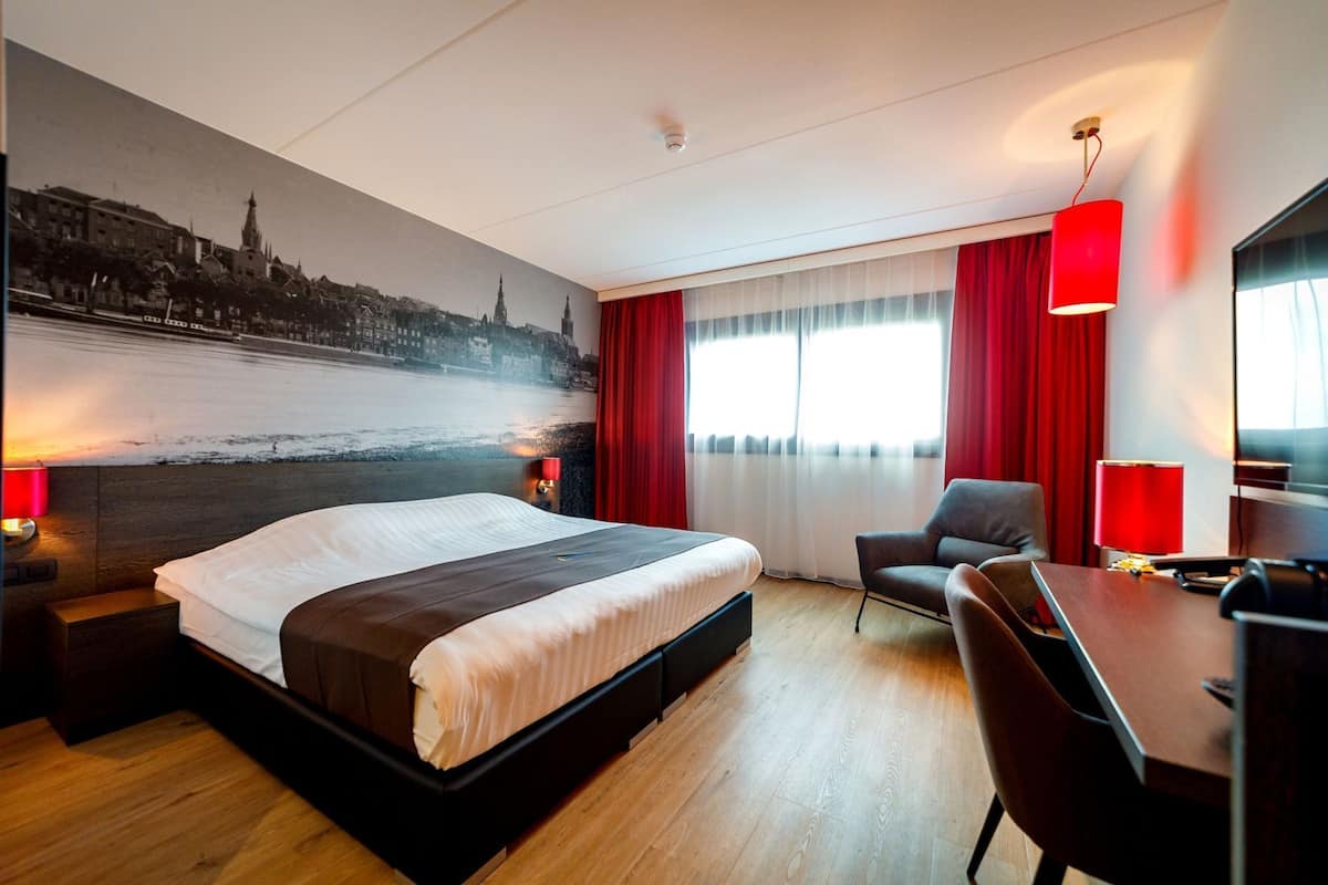 Une chambre d’hôtel avec un grand lit, un bureau, une chaise, une lampe de bureau et une fresque représentant un paysage urbain sur le mur.