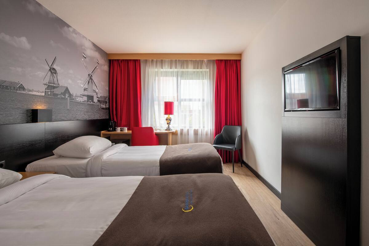 A hotel room with two beds, a desk, a chair, a TV, and a mural of windmills.