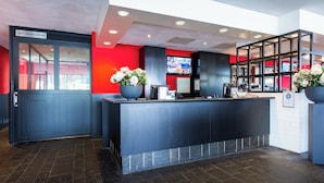 Un comptoir d’accueil avec un plan de travail foncé, une porte en verre portant l’inscription « salle de jeux / fumoir », et un mur rouge avec un écran de télévision en arrière-plan.