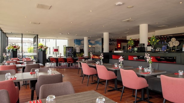 Un restaurant moderne doté de plusieurs espaces de restauration, notamment un bar et un coin salon avec des chaises rouges. Les tables sont dressées avec de la verrerie et des serviettes.