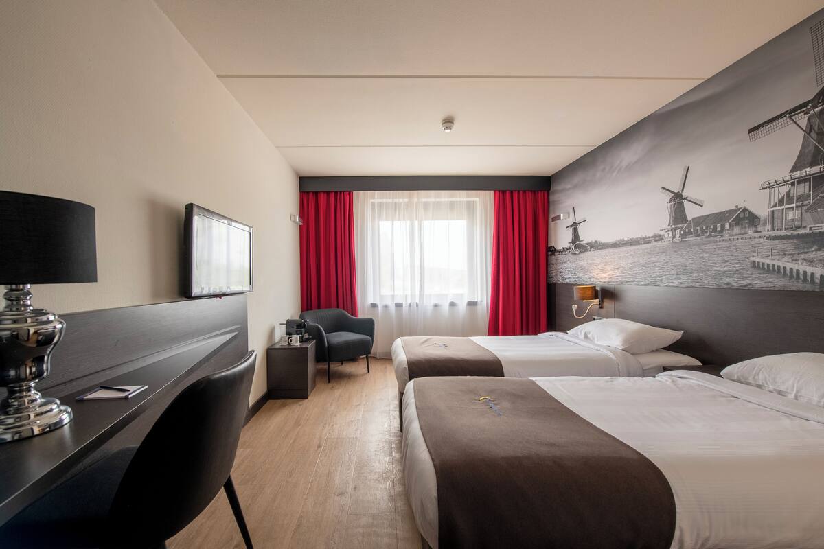 A hotel room with two beds, a desk, a chair, a TV, and a large window with red curtains.