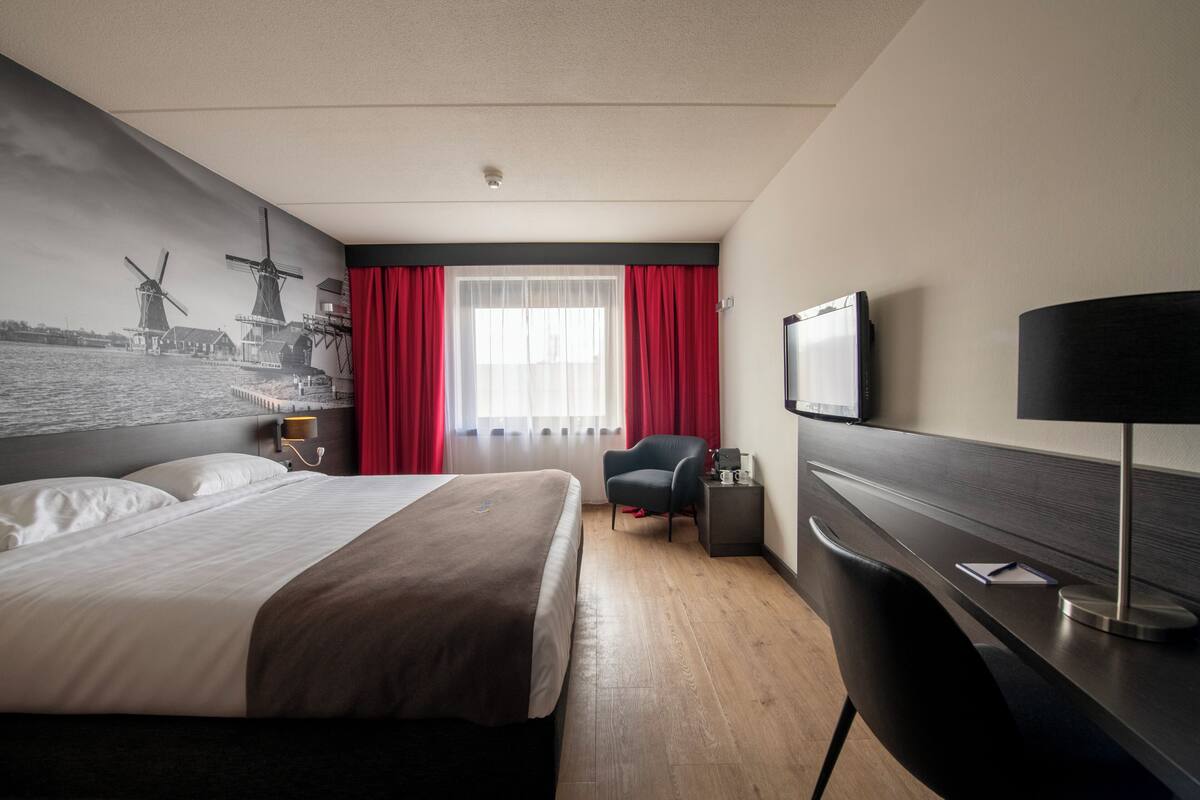 A hotel room with a bed, a desk with a lamp, a chair, a television, and a window with red curtains.