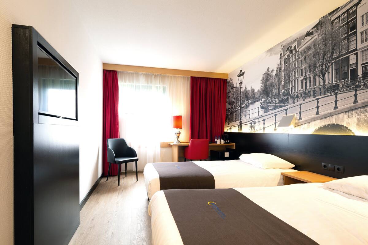 A hotel room with two beds, a desk, a chair, and a large window with red curtains.