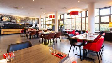 A modern restaurant with high tables and red chairs, large windows, and a mural on the wall.