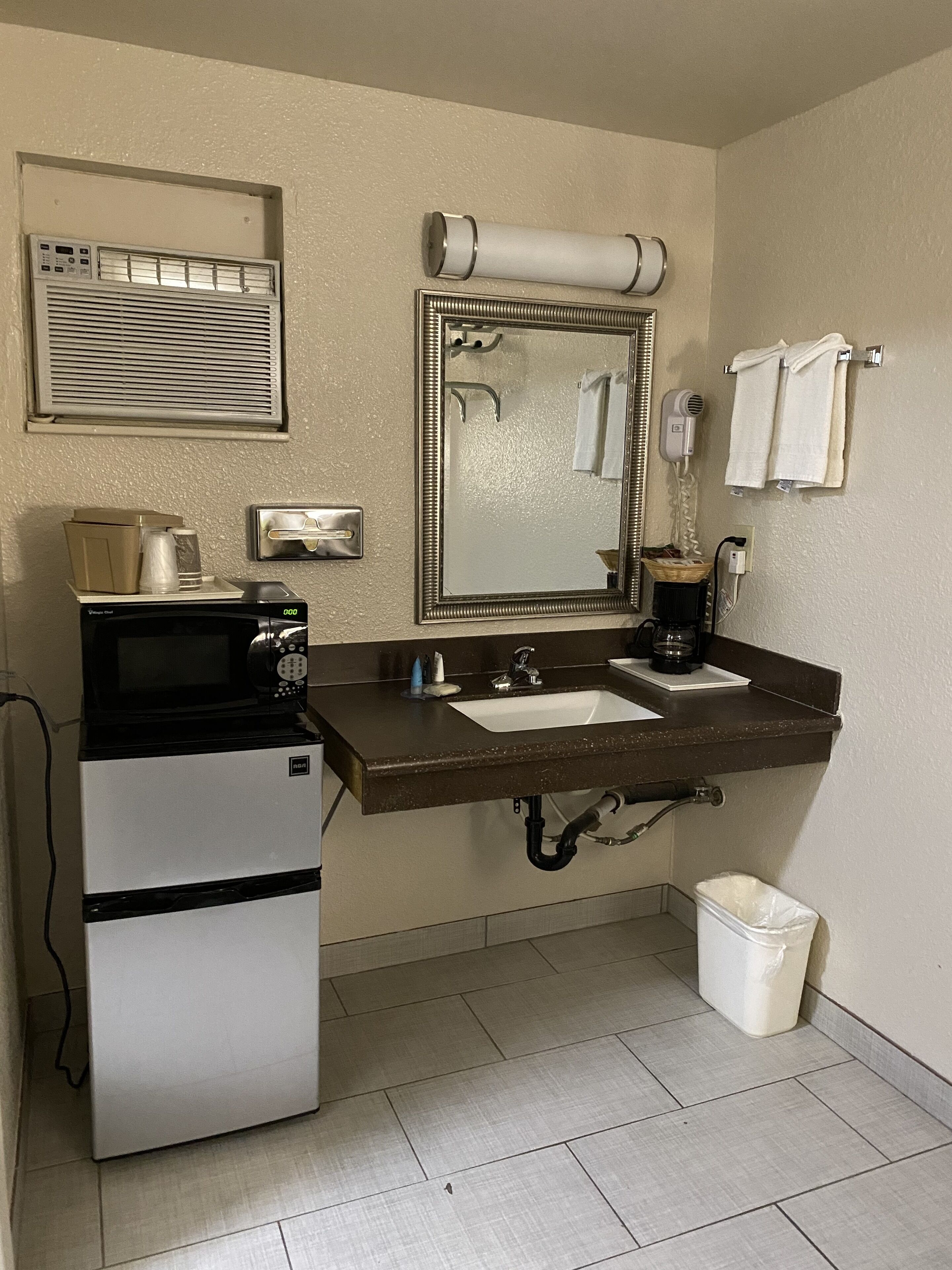 Een kleine badkamer met een wastafel, spiegel, handdoekhouder, haardroger en een klein prullenbakje.