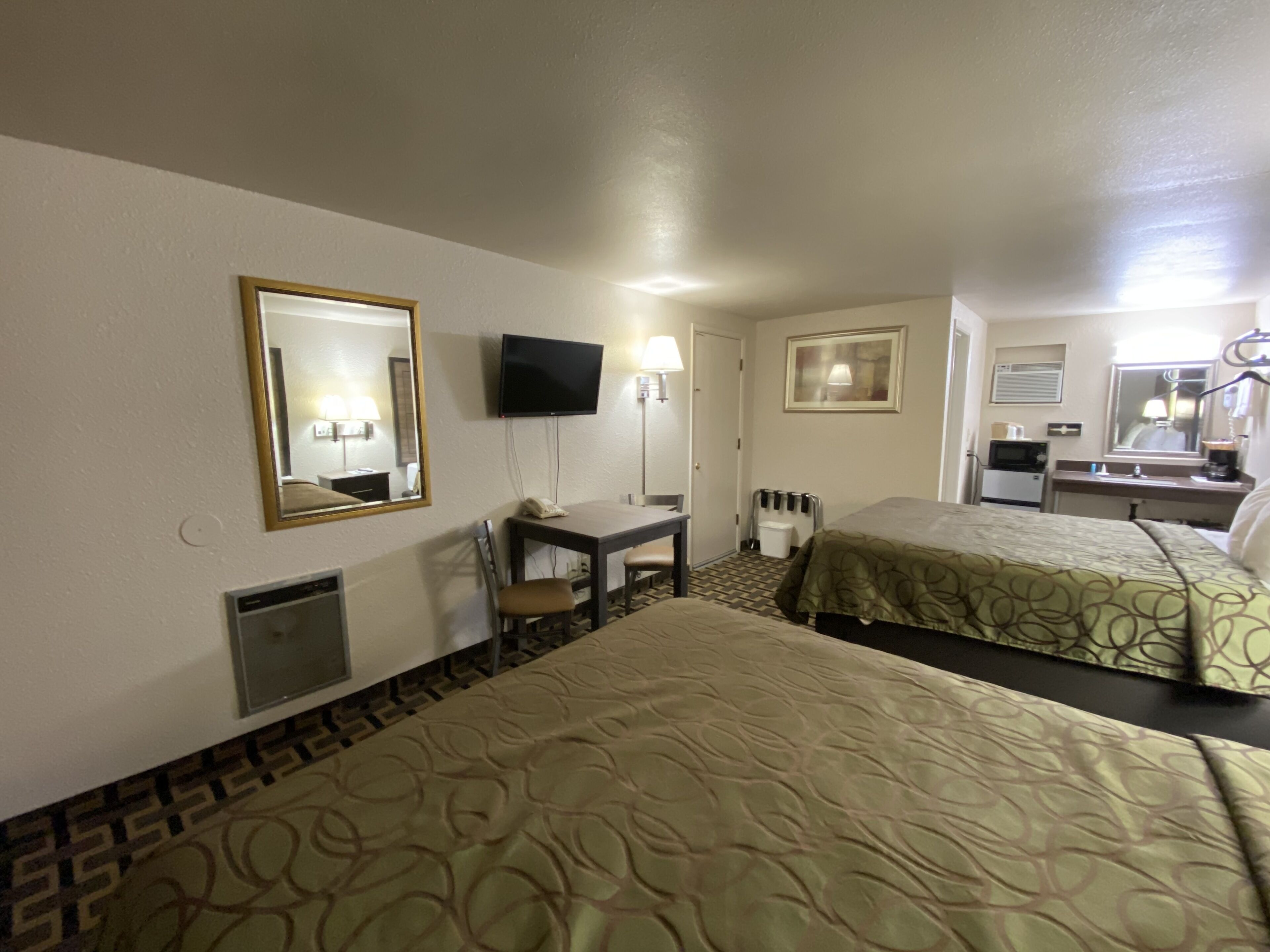 A hotel room with two beds, a desk with a chair, a television, a mirror, and a small kitchenette.