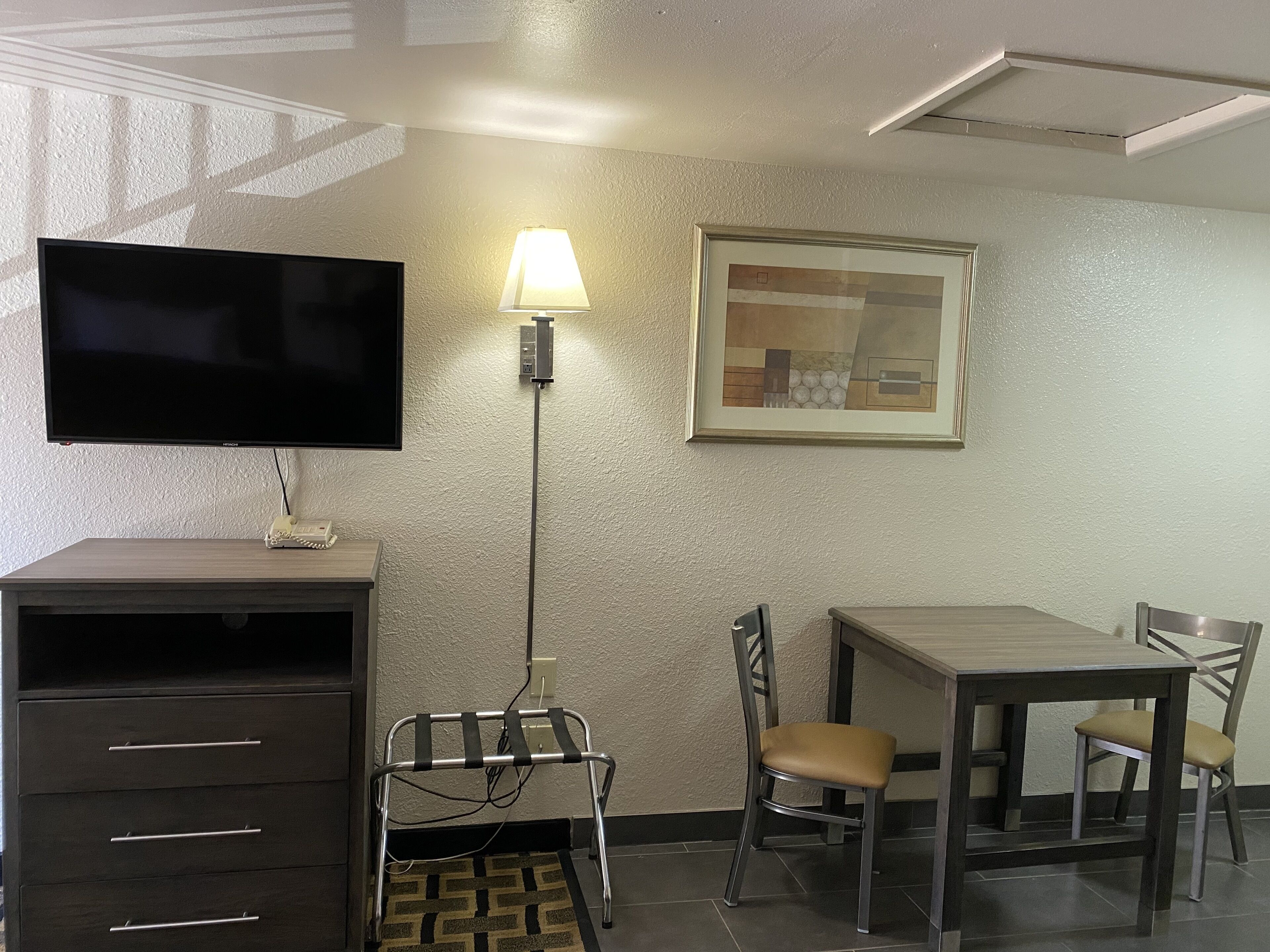 Een hotelkamer met een flatscreen televisie, een bureau, een stoel, een lamp en een ingelijst kunstwerk aan de muur.
