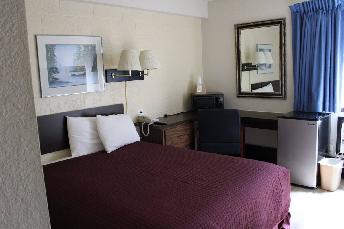 Ein Hotelzimmer mit einem großen Bett, einem Schreibtisch mit Telefon, einem Fernseher, einem Spiegel und einem Mülleimer.