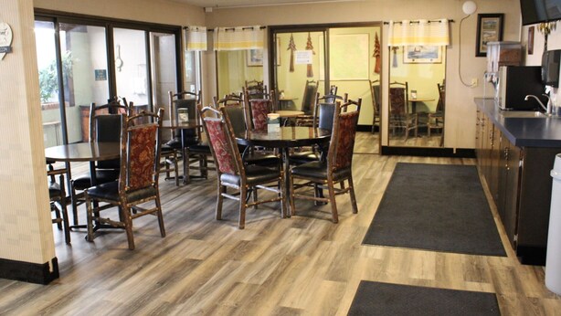 A dining area with wooden tables and chairs, a kitchen area with a sink and cabinets, and a view of the outside through large windows. - Aero Inn (Kalispell)