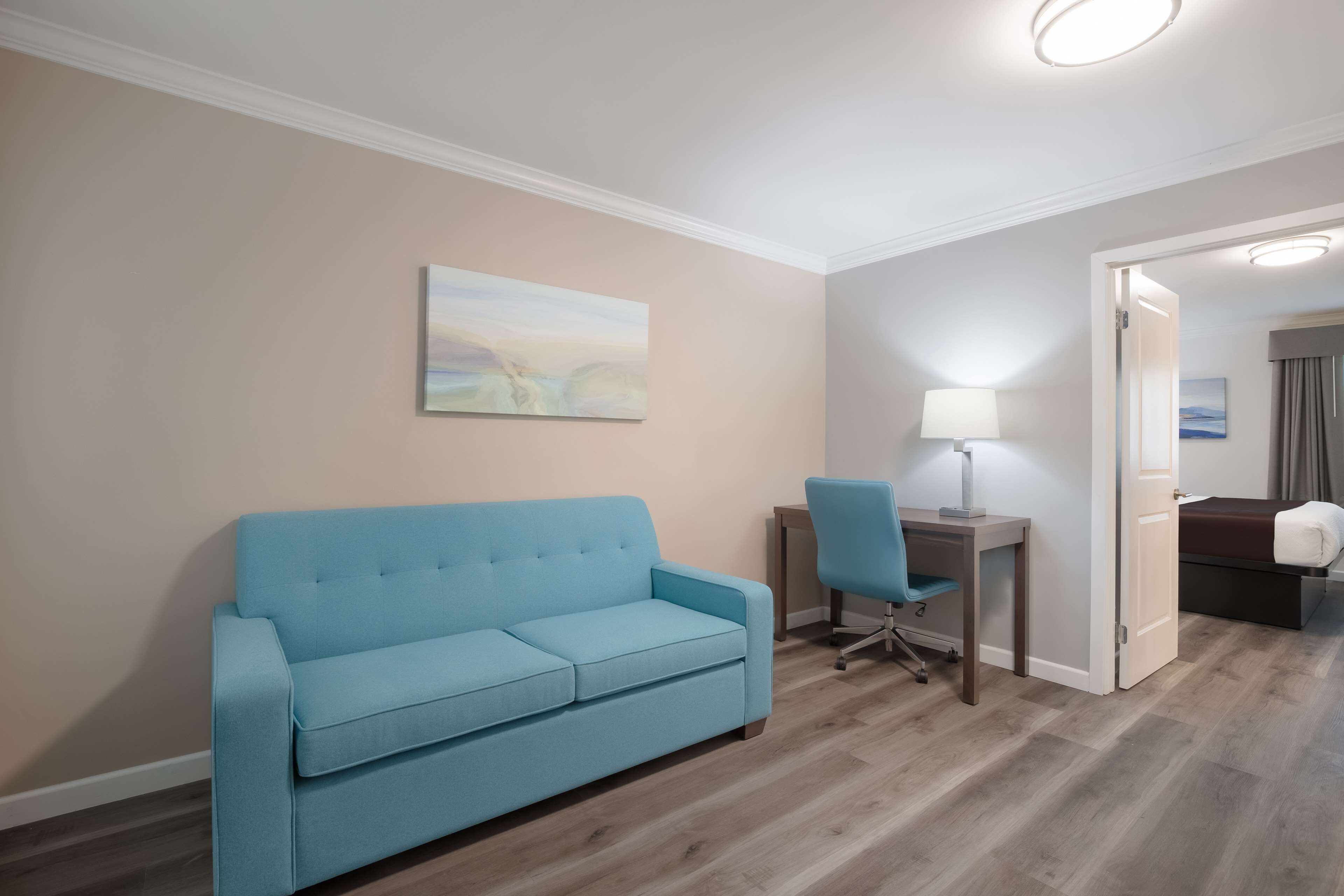 A room with a blue sofa, a desk with a chair, a lamp, and a bed with a brown headboard.