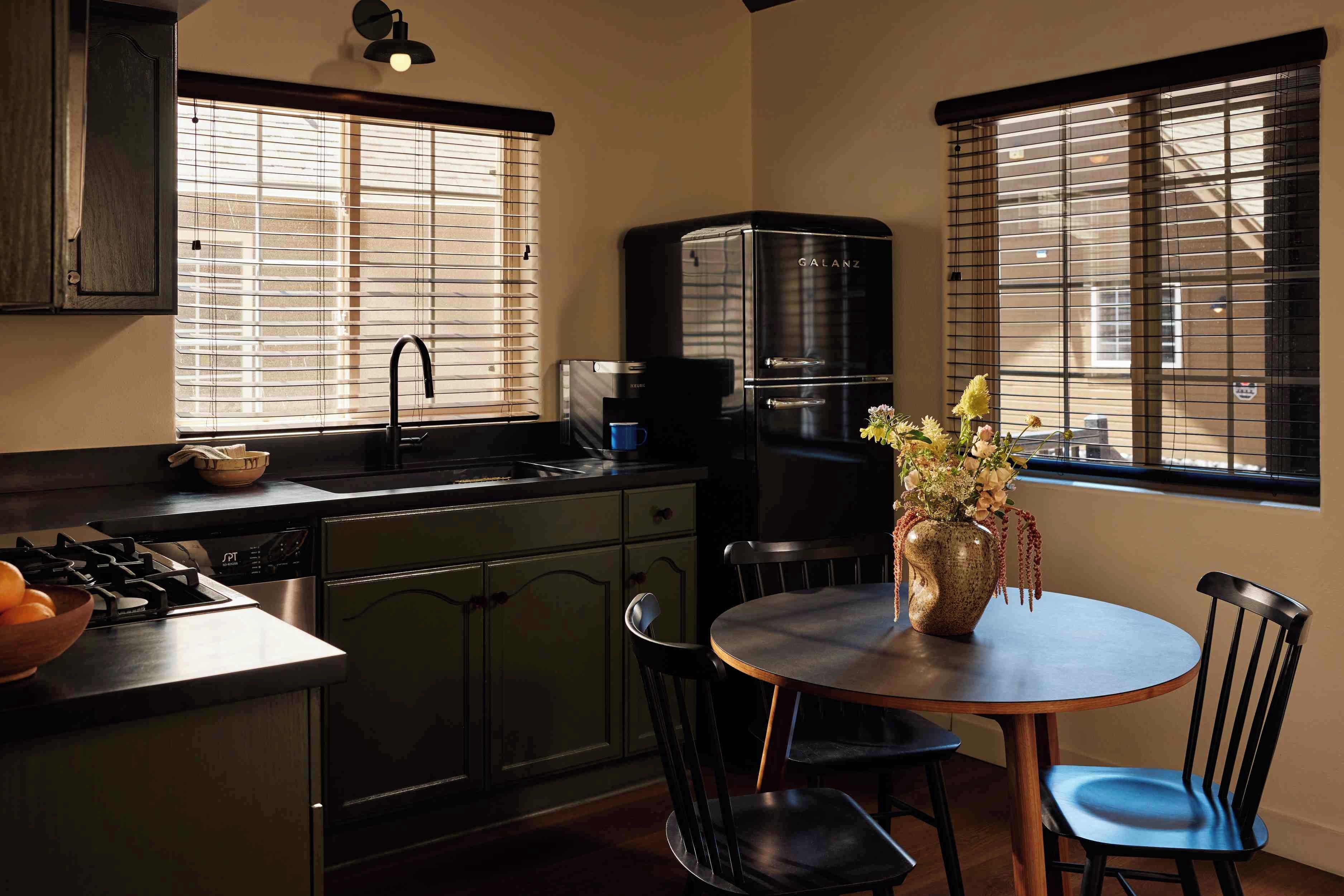 A kitchen with a stove, sink, and a coffee maker. There is a dining table with chairs and a vase with flowers.