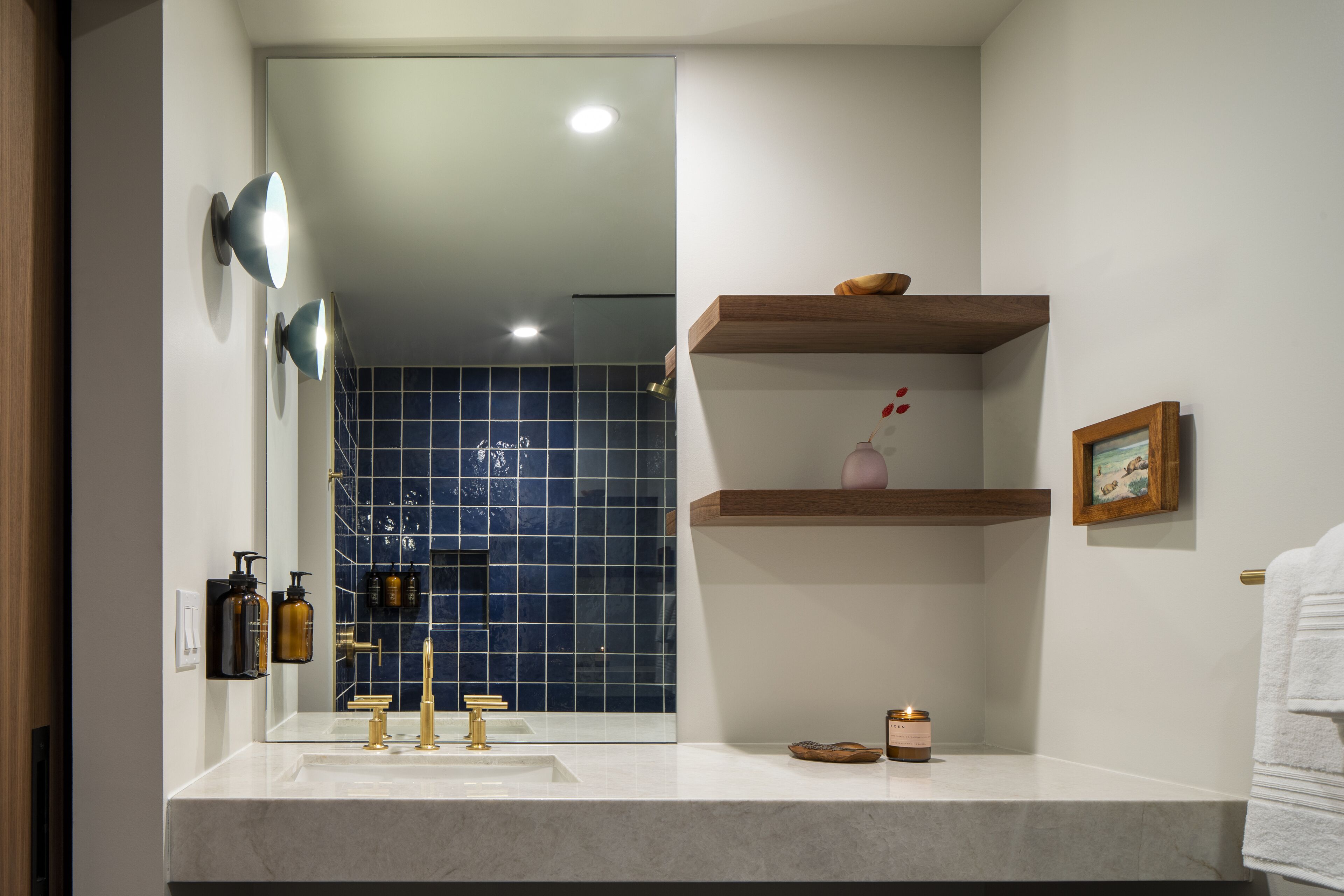 A bathroom with a white sink, gold faucets, and a mirror. There are two wall-mounted lights, a shelf with a vase and a picture frame, and a candle on the counter.