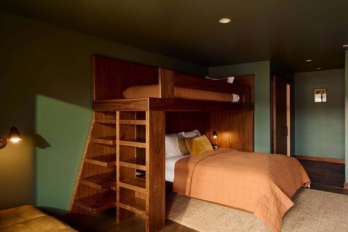 A bunk bed with a wooden ladder, a bedside table with a lamp, and a wall-mounted shelf.
