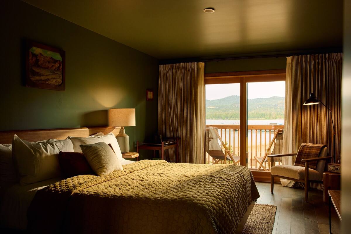 A hotel room with a large bed, a view of the outdoors, a chair, and a lamp.
