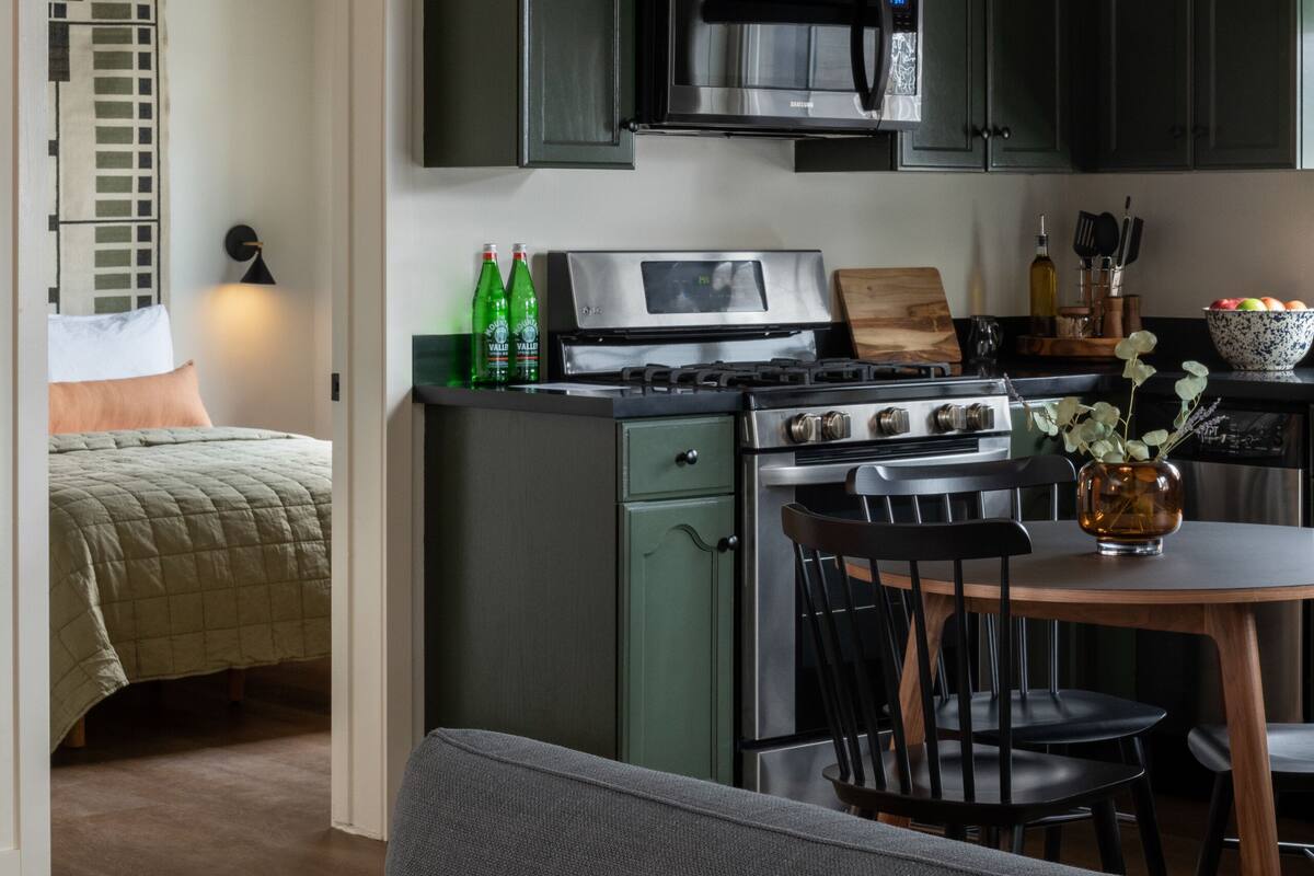 A compact kitchen with dark green cabinets, a stainless steel stove, and a dining area with a table and chairs.