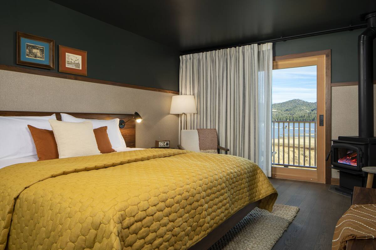 A hotel room with a large bed, a nightstand with a lamp, a chair, a view of the outdoors through a sliding door, and a fireplace.
