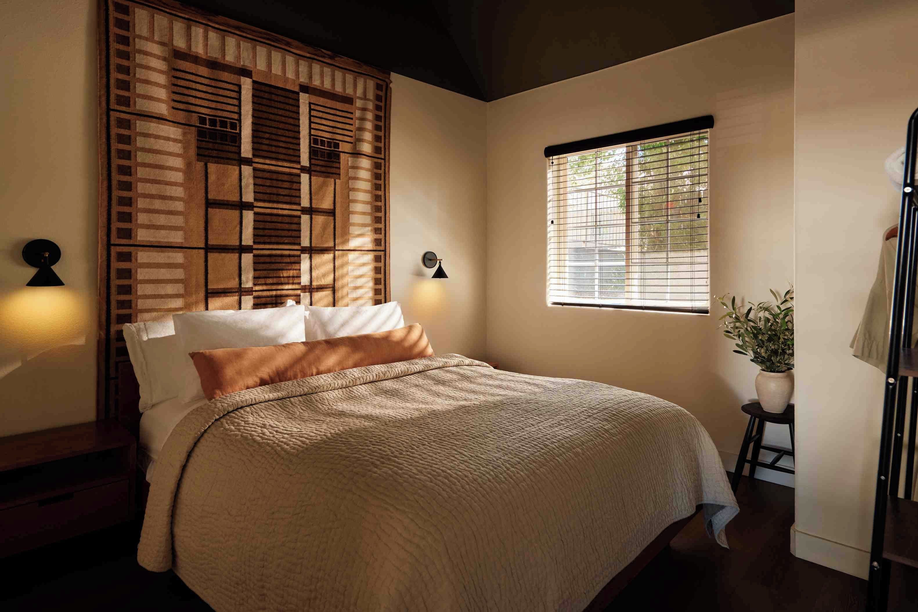 Un dormitorio con cama, ventana con persianas, un reloj en la pared y una planta en maceta.