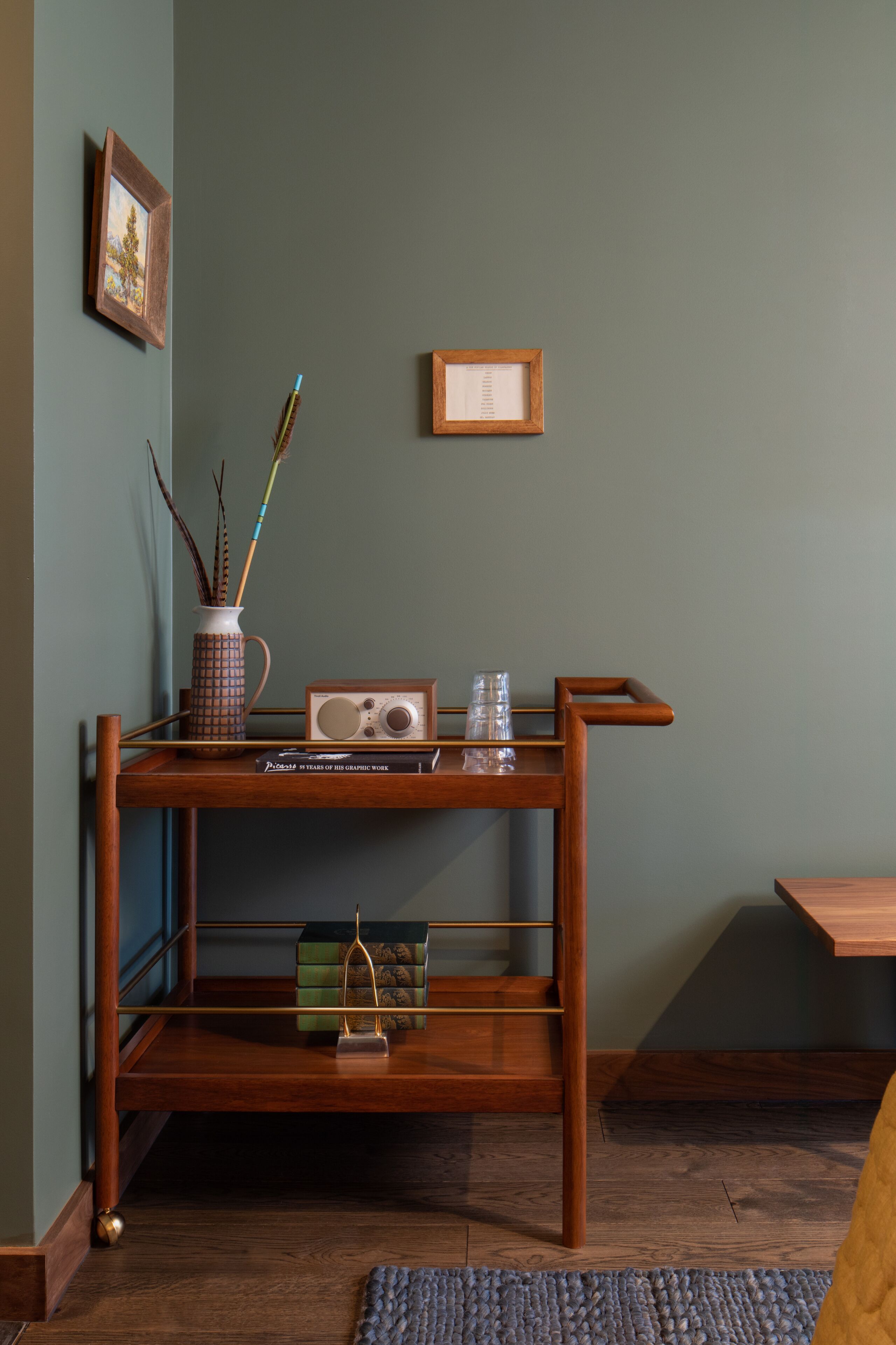 Una consola de madera con una radio, un florero con ramas y un cuadro en una pared verde.