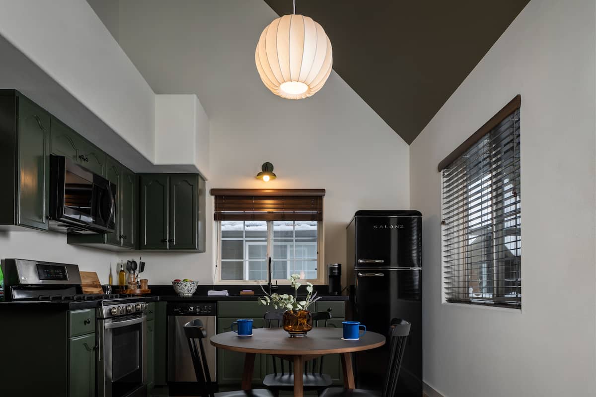 A compact kitchen with green cabinets, a black refrigerator, a wooden dining table with chairs, and a hanging light fixture.