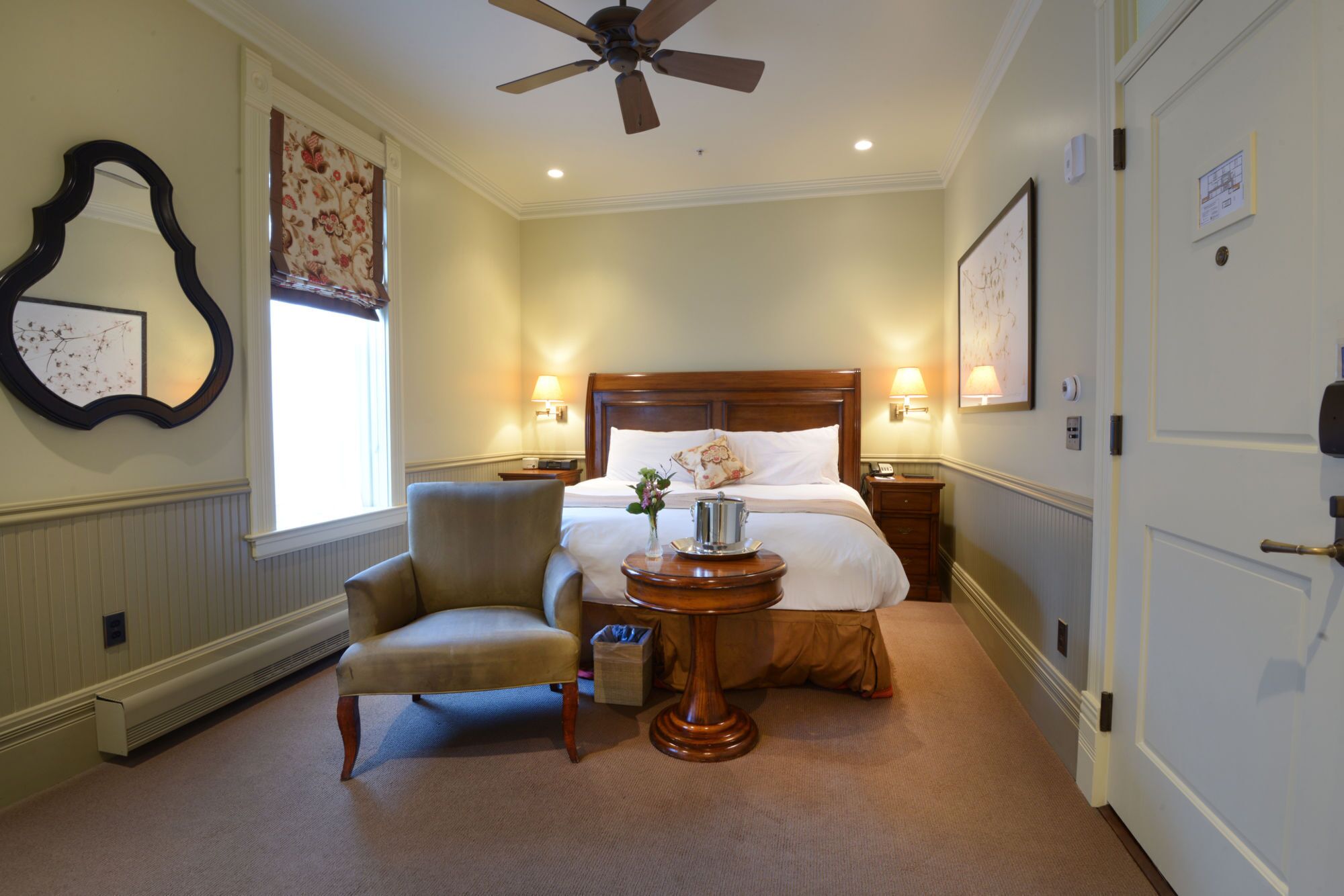 A hotel room with a bed, a chair, a small table with a lamp, and a ceiling fan.