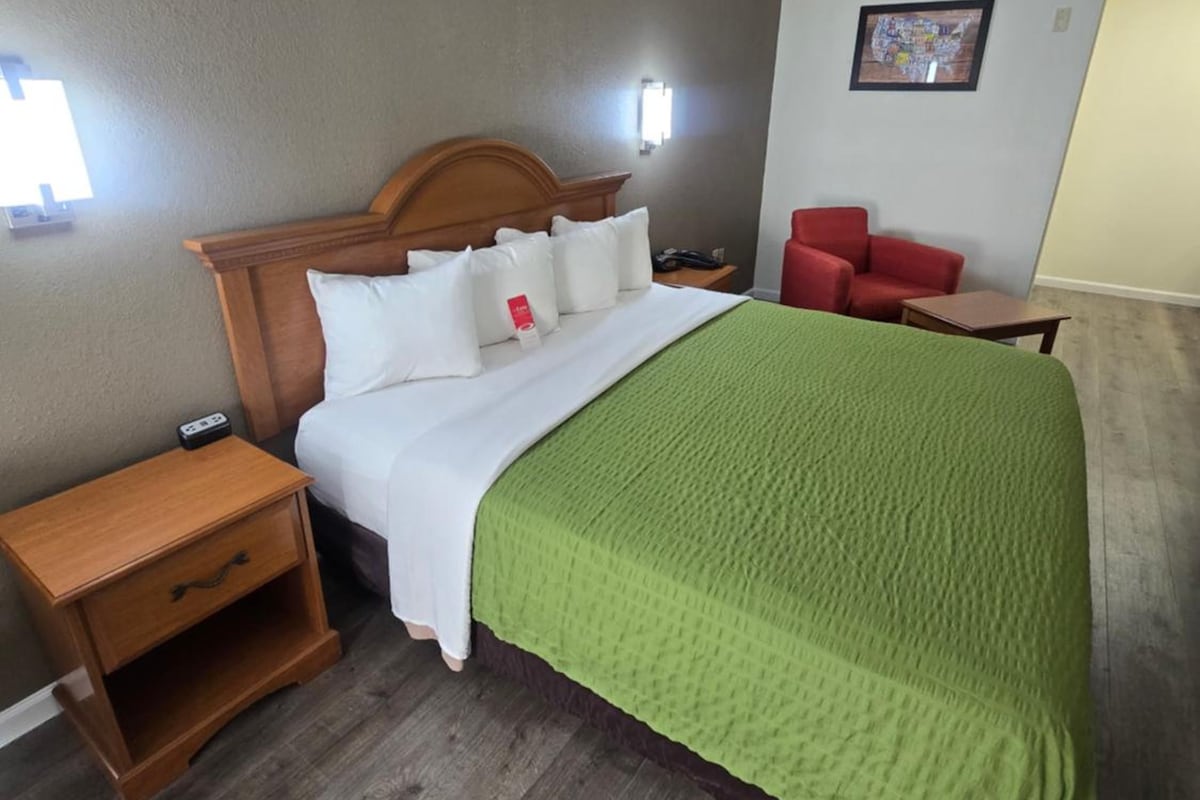 A hotel room with a bed, a nightstand, a red chair, and a wall-mounted light.