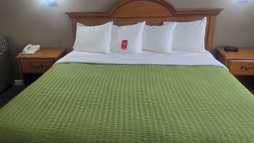 Una habitación de hotel con una cama grande, un cabecero de madera, una colcha de textura verde, almohadas blancas, una mesita de noche con un teléfono y un letrero rojo de Easy hotel.