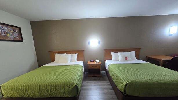 A hotel room with two beds, each with a green bedspread and white pillows. There is a nightstand with a phone and a lamp between the beds. The room has a wooden floor and a painting on the wall.