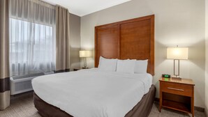 A hotel room with a large bed, two bedside lamps, a wooden headboard, a nightstand with a lamp, and a window with curtains. - Comfort Suites Brenham (Brenham)