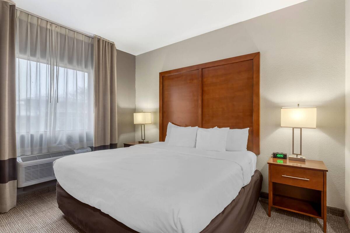 Habitación de hotel con una cama grande, dos lámparas de noche, un cabecero de madera, una mesita de noche con lámpara y una ventana con cortinas.