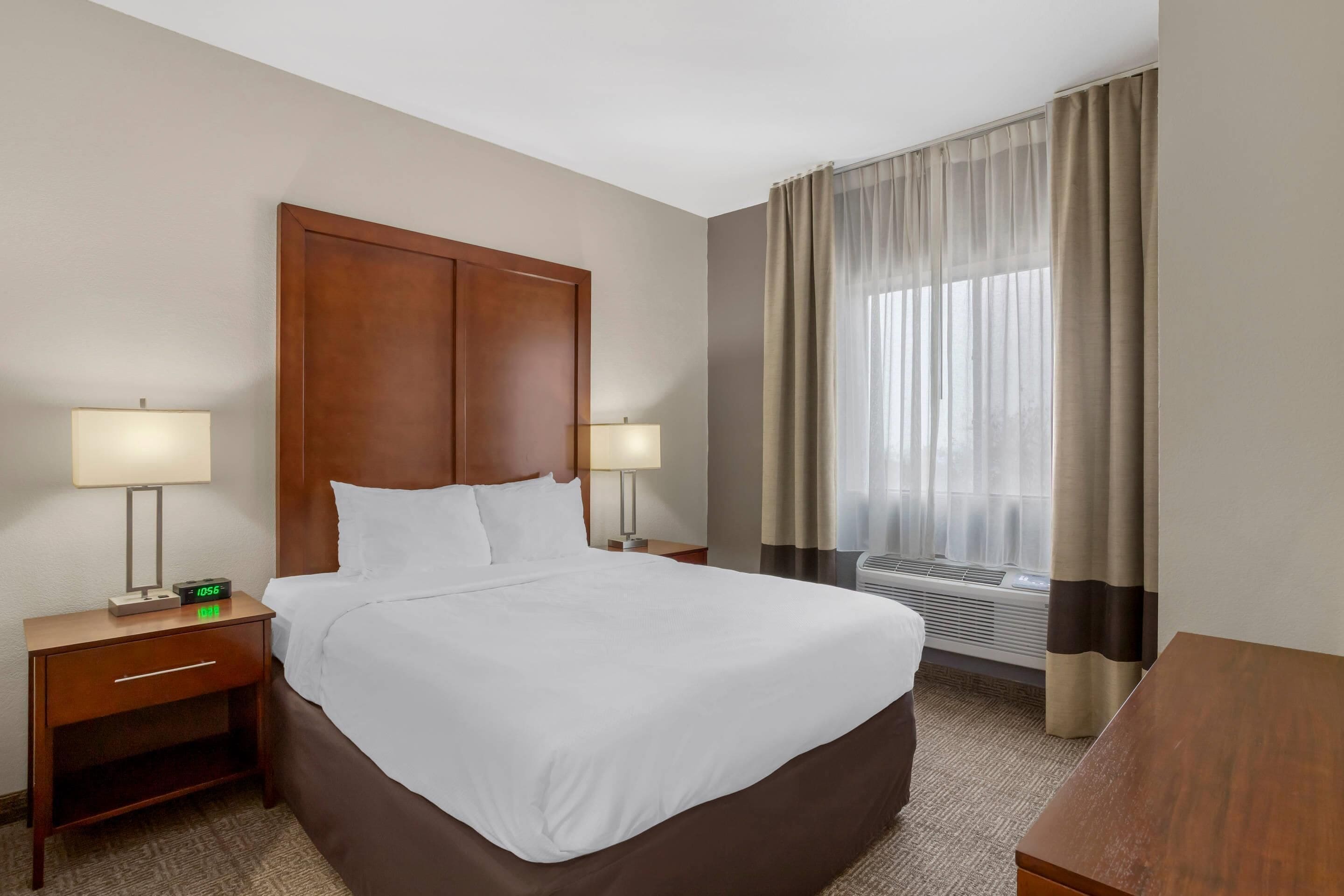 A hotel room with a large bed, two bedside tables with lamps, a window with curtains, and an air conditioning unit.