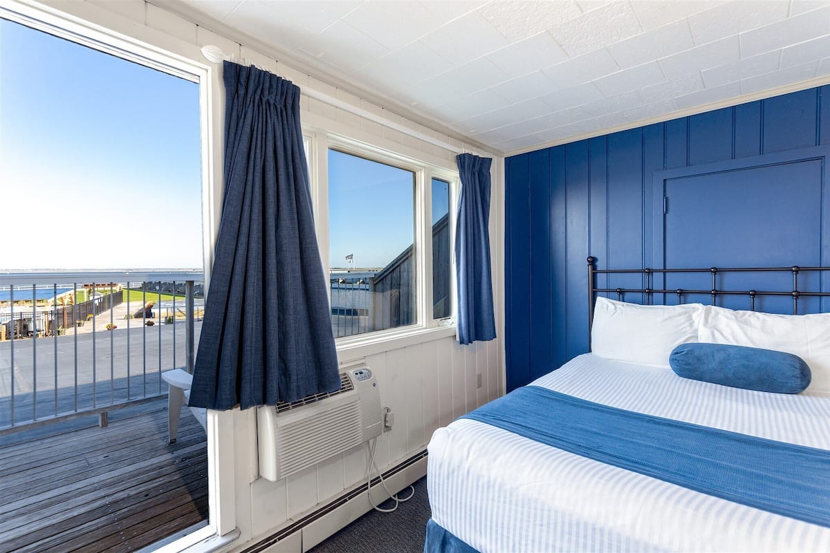 Une chambre avec un lit, une fenêtre avec des rideaux, une vue sur la mer et un balcon avec une balustrade.
