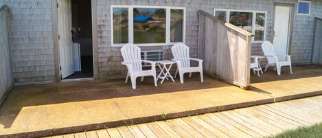 Un patio techado con dos sillas Adirondack blancas y una mesita, una terraza de madera, y una casa con revestimiento gris y techo azul.