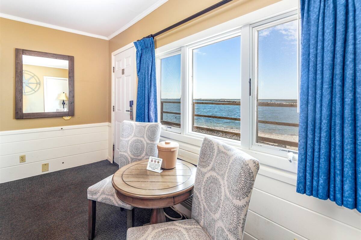 Une pièce avec vue sur la mer, une table ronde sur laquelle est posé un panneau, des chaises à motifs et un grand miroir au mur.