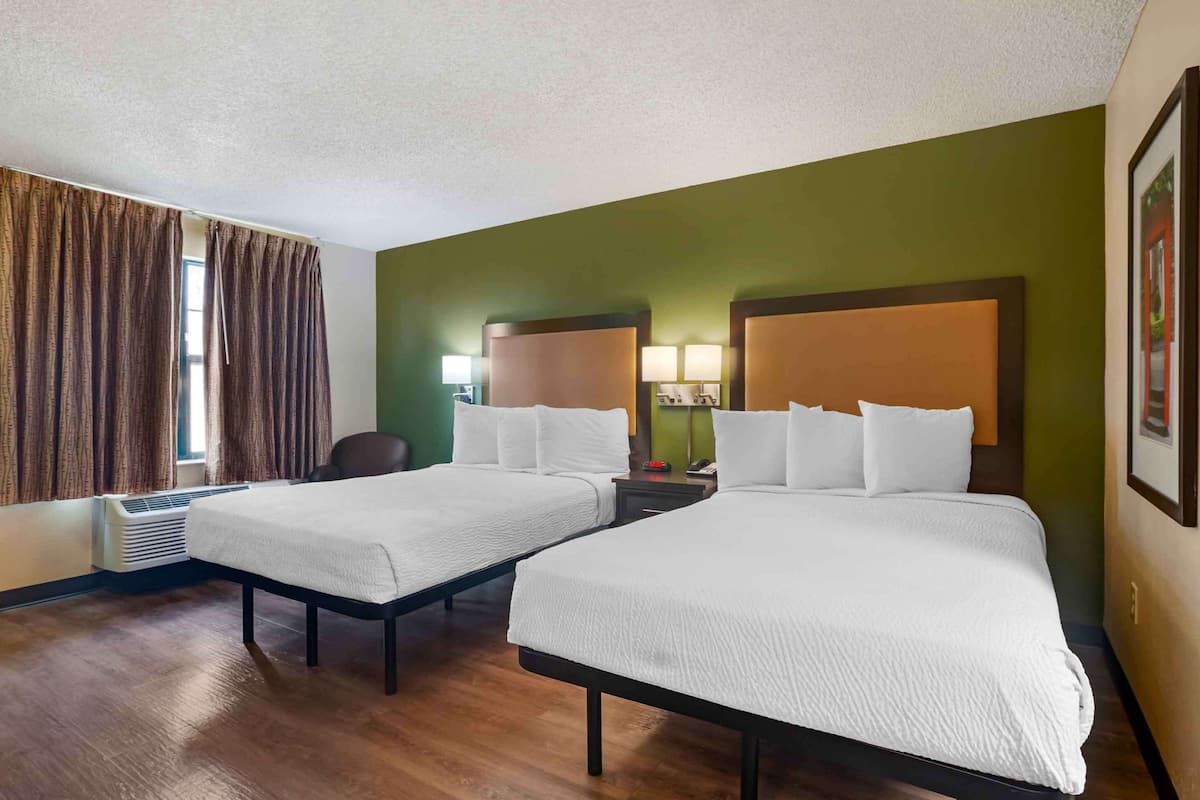 A hotel room with two beds, a chair, a desk with a lamp, and a framed picture on the wall.